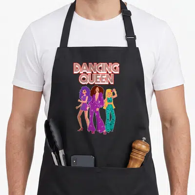 Queen 1970s apron - DANCING QUEEN text above three cartoon women in purple and teal outfits — shown on a black cotton canvas apron against a white background