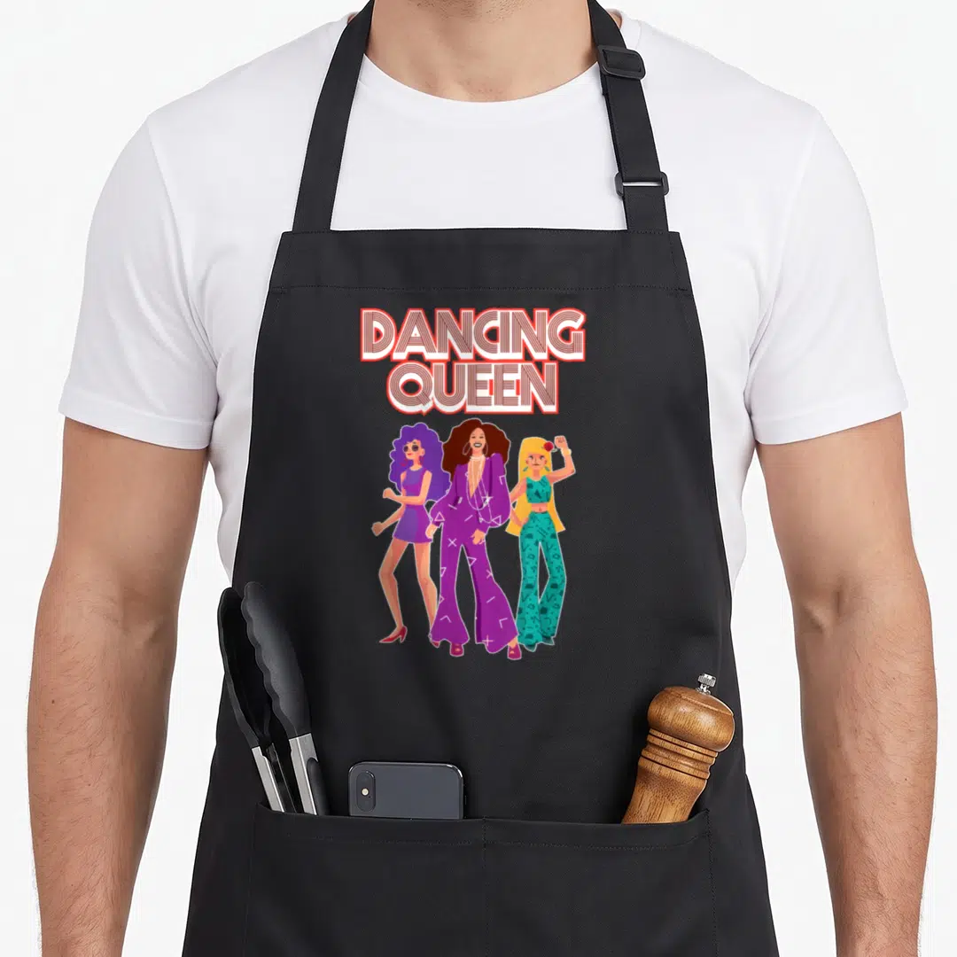 Queen 1970s apron - DANCING QUEEN text above three cartoon women in purple and teal outfits — shown on a black cotton canvas apron against a white background