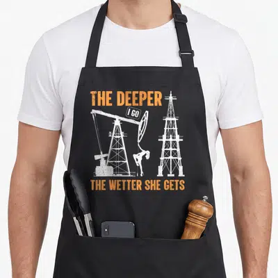 Oil lifestyle apron - Distressed orange text reading THE DEEPER I GO THE WETTER SHE GETS above white oil rig and woman — shown on a black cotton canvas apron against a white background