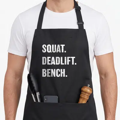Weightlifting bench apron - White SQUAT. DEADLIFT. BENCH. stacked text on black background — shown on a black cotton canvas apron against a white background