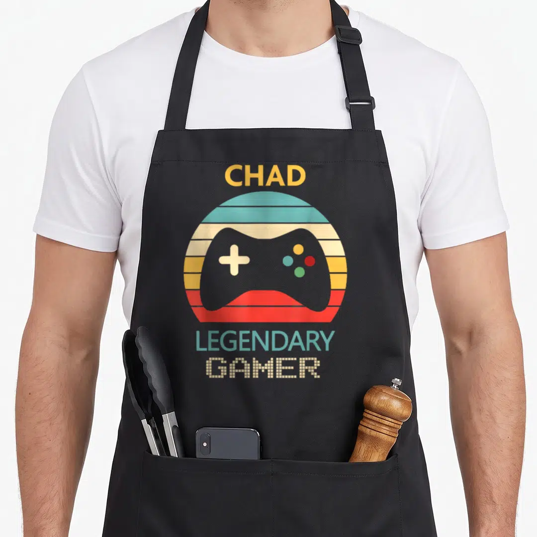 Legendary chad apron - Retro game controller in striped circle with CHAD LEGENDARY GAMER text — shown on a black cotton canvas apron against a white background