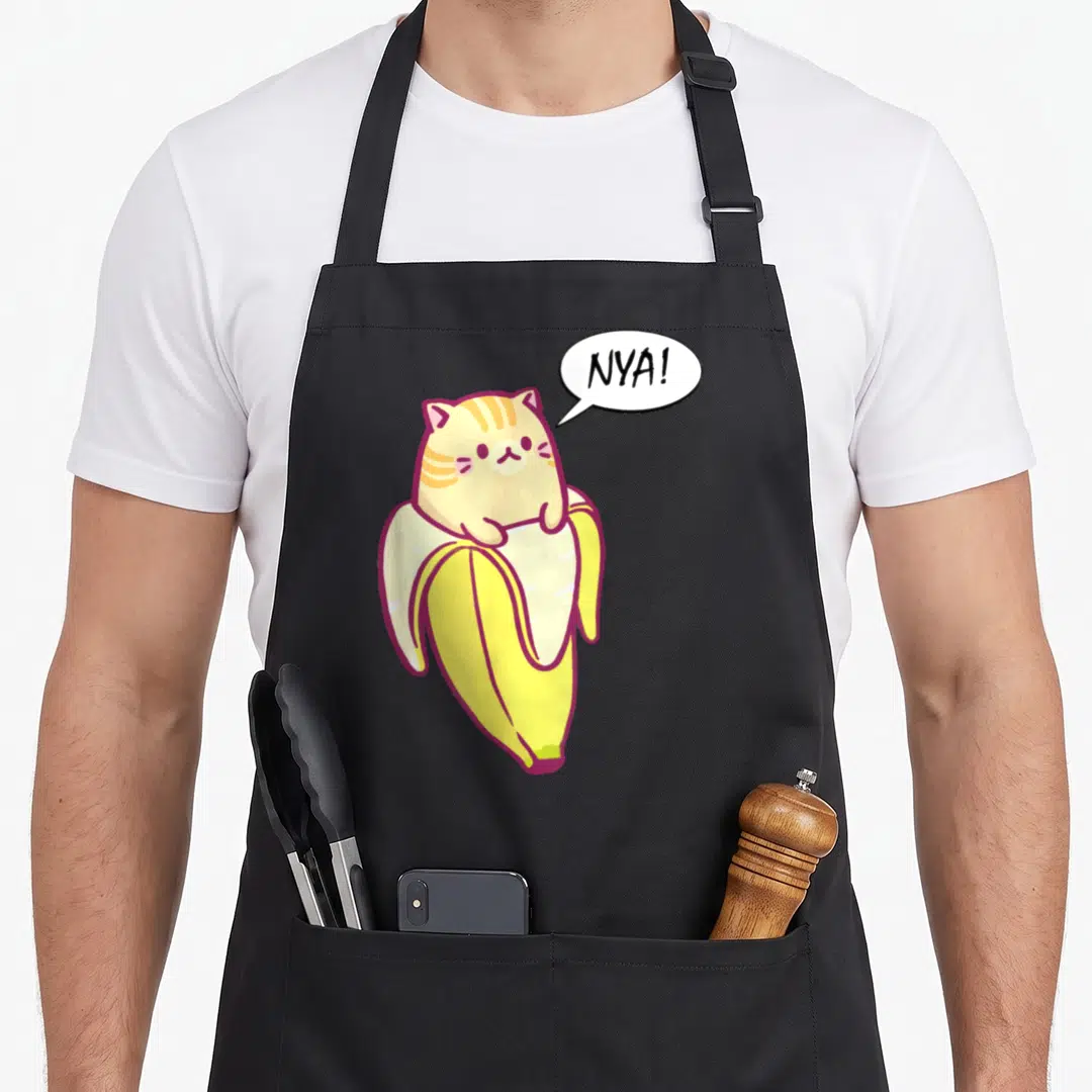 Bananya banana apron - Cartoon cream cat sitting in a yellow peeled banana with a white speech bubble reading NYA — shown on a black cotton canvas apron against a white background