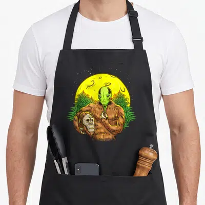 Solved bigfoot apron - Green alien head in brown bigfoot suit under yellow moon and two pine trees — shown on a black cotton canvas apron against a white background