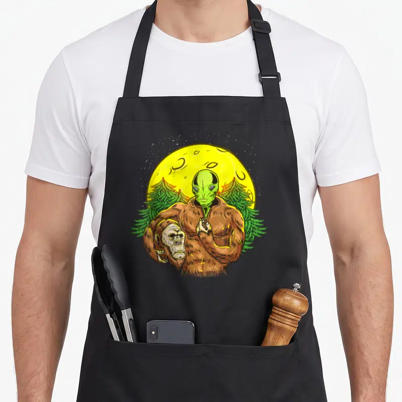 Solved bigfoot apron - Green alien head in brown bigfoot suit under yellow moon and two pine trees — shown on a black cotton canvas apron against a white background