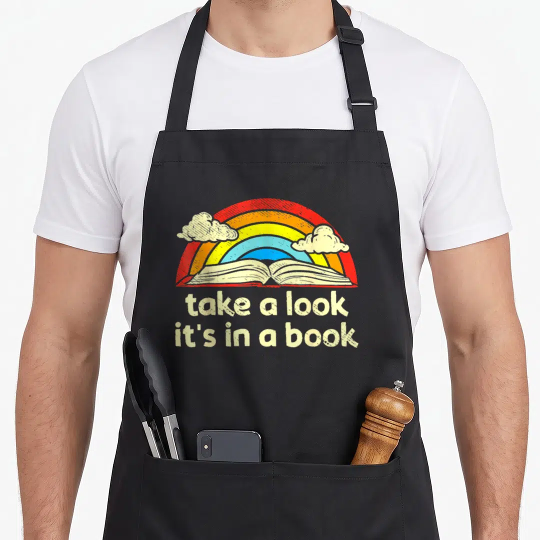 Book reading rainbow apron - Cartoon open book under rainbow arch with two clouds and stacked text take a look it's — shown on a black cotton canvas apron against a white background