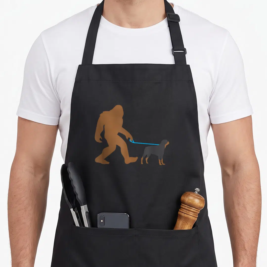 Rottweiler walking apron - Brown Bigfoot silhouette holding blue leash attached to dark grey Rottweiler dog standing — shown on a black cotton canvas apron against a white background