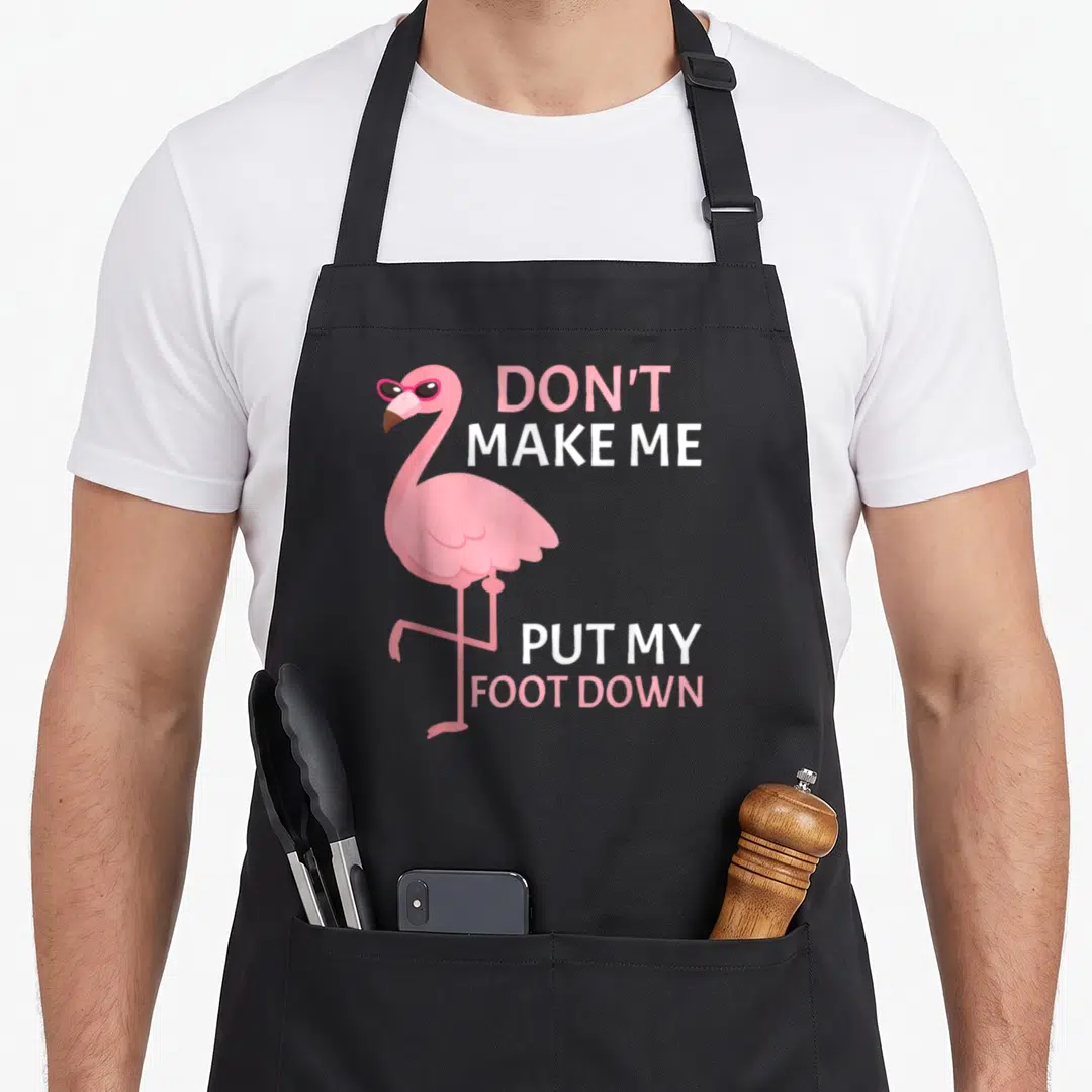 Foot pink flamingo apron for mom - Pink cartoon flamingo with sunglasses beside stacked text DON'T MAKE ME PUT MY FOOT — shown on a black cotton canvas apron against a white background