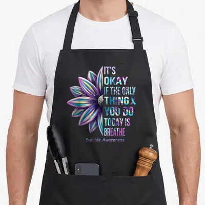 Okay depression awareness apron - Colorful watercolor flower next to stacked text IT'S OKAY IF THE ONLY THING YOU DO — shown on a black cotton canvas apron against a white background