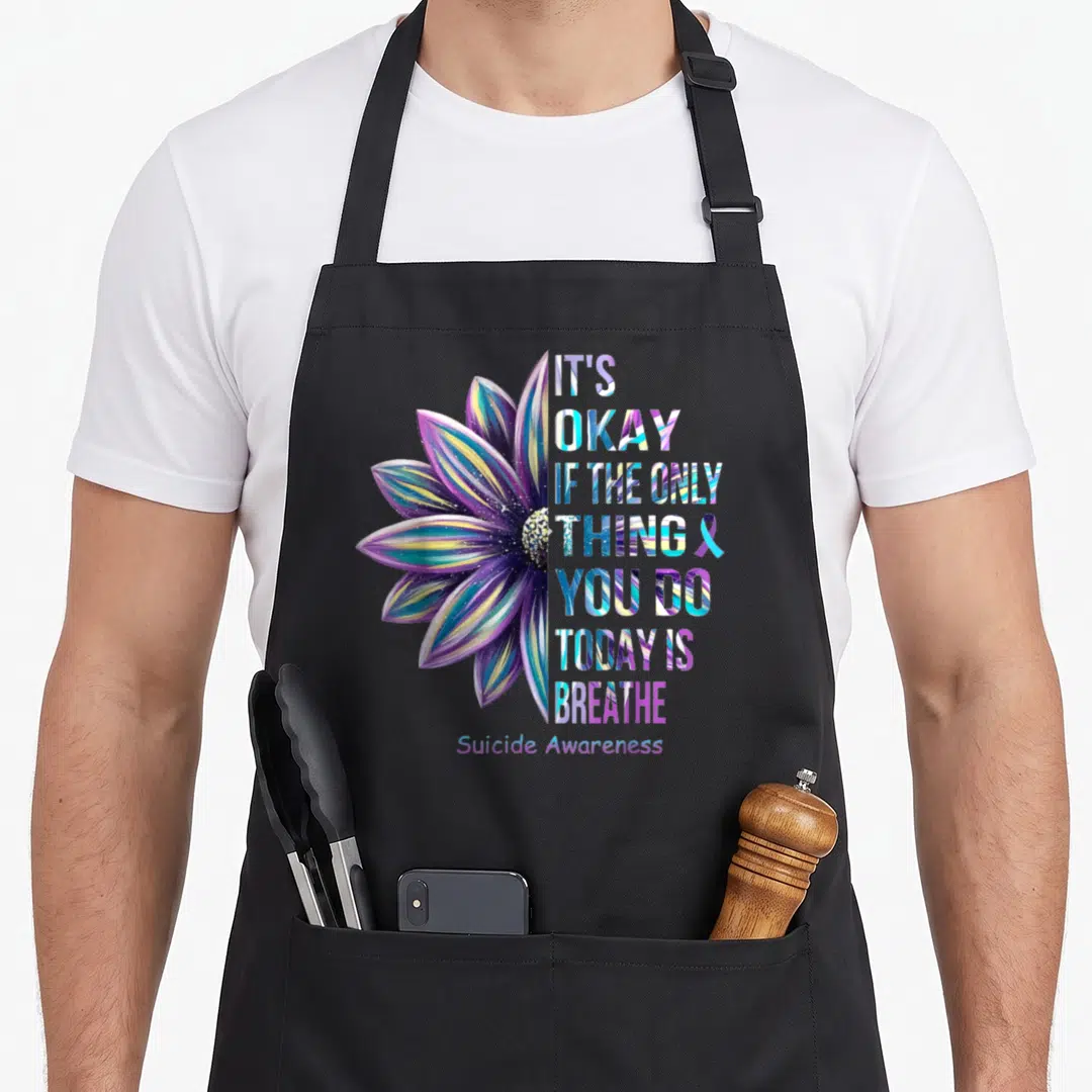 Okay depression awareness apron - Colorful watercolor flower next to stacked text IT'S OKAY IF THE ONLY THING YOU DO — shown on a black cotton canvas apron against a white background