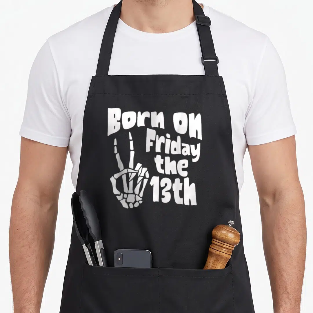 Funny halloween apron - White skeleton hand peace sign next to Born On Friday the 13th text — shown on a black cotton canvas apron against a white background
