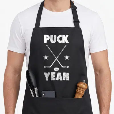 Puck goblin apron - White distressed PUCK YEAH text with crossed hockey sticks and puck on black background — shown on a black cotton canvas apron against a white background