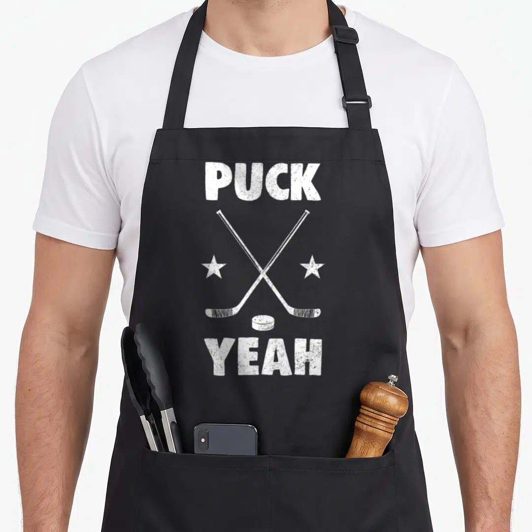 Puck goblin apron - White distressed PUCK YEAH text with crossed hockey sticks and puck on black background — shown on a black cotton canvas apron against a white background