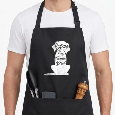 Rescue top dog aprons - Distressed white dog silhouette containing stacked black text reading Rescue Is My Favorite — shown on a black cotton canvas apron against a white background