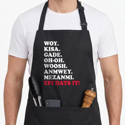 Cute haitian pride apron - Stacked white and red Haitian Creole words Woy Kisa Gade Epi Dats It — shown on a black cotton canvas apron against a white background