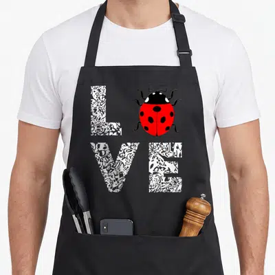 Entomology ladybug apron - Red ladybug replaces letter O in textured white LOVE text — shown on a black cotton canvas apron against a white background
