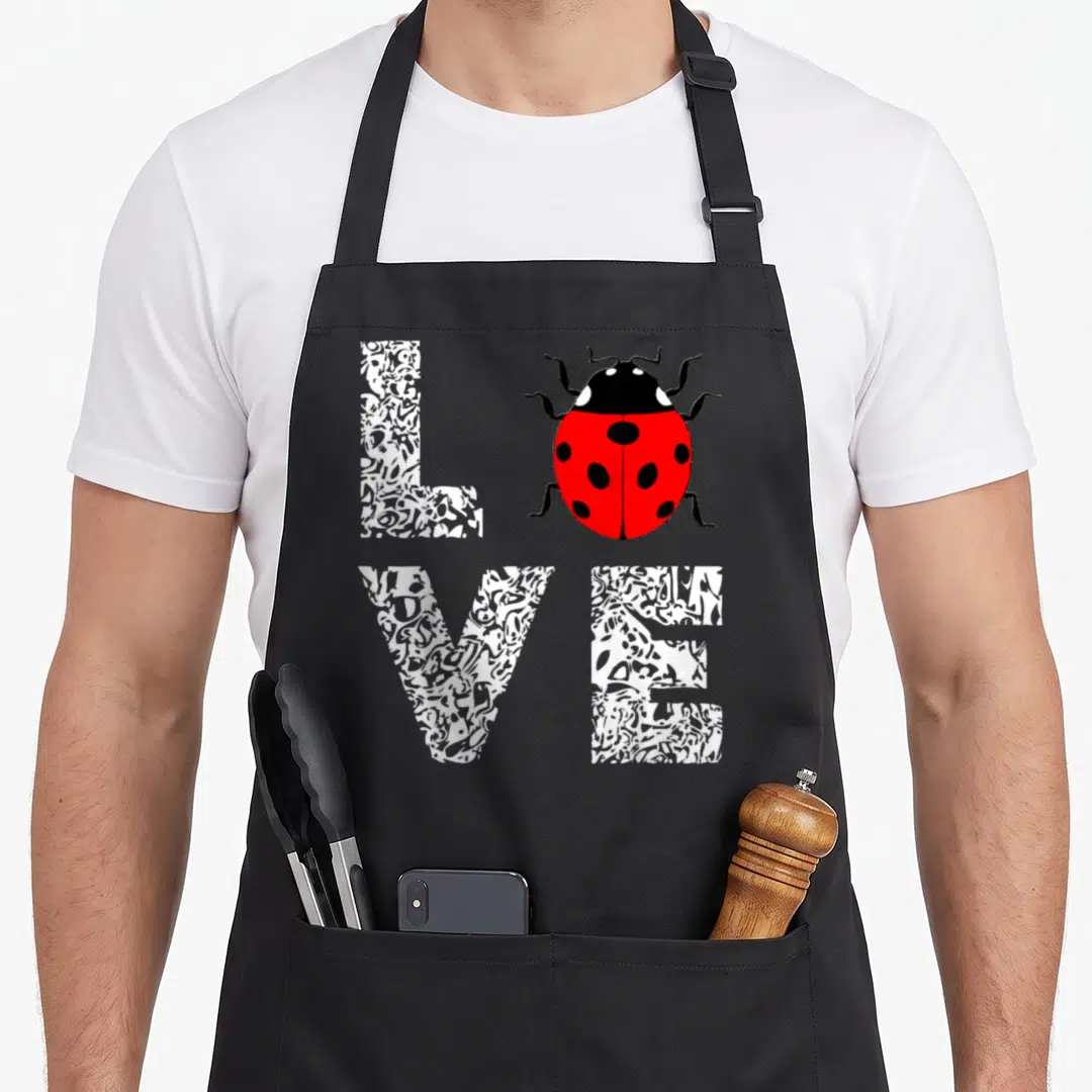 Entomology ladybug apron - Red ladybug replaces letter O in textured white LOVE text — shown on a black cotton canvas apron against a white background