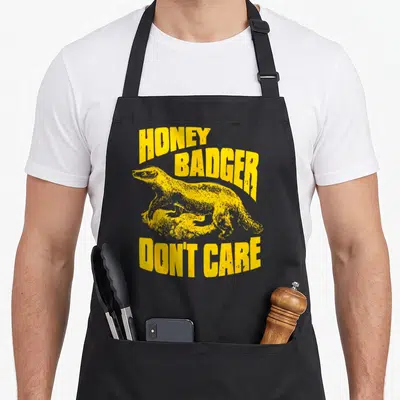 Care badger apron - Yellow distressed text reading HONEY BADGER DON'T CARE around a badger on a rock — shown on a black cotton canvas apron against a white background