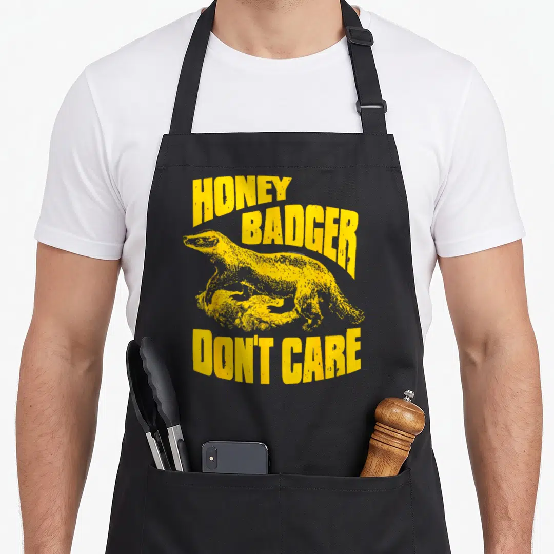Care badger apron - Yellow distressed text reading HONEY BADGER DON'T CARE around a badger on a rock — shown on a black cotton canvas apron against a white background