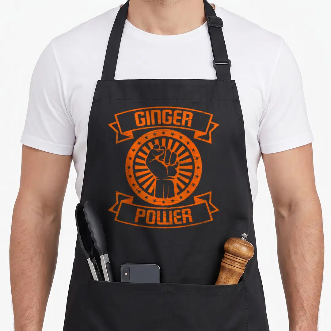 Ginger hair apron - Orange GINGER POWER text with raised fist inside sunburst circle and ribbons — shown on a black cotton canvas apron against a white background