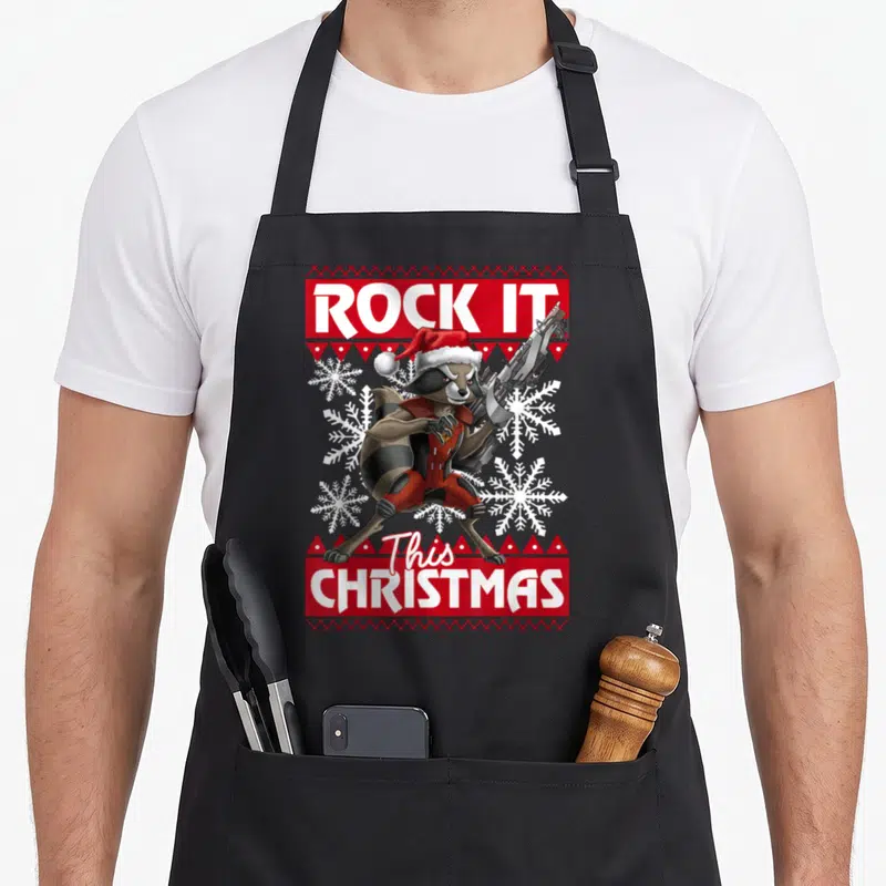 Rocket christmas apron - Cartoon raccoon in santa hat holding gun with red knitted border and snowflakes — shown on a black cotton canvas apron against a white background