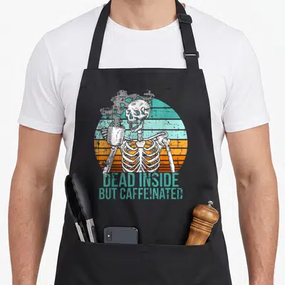 Dead coffee aprons - Skeleton holding mug in front of striped sun with teal text reading DEAD INSIDE BUT CAFFEINATED — shown on a black cotton canvas apron against a white background