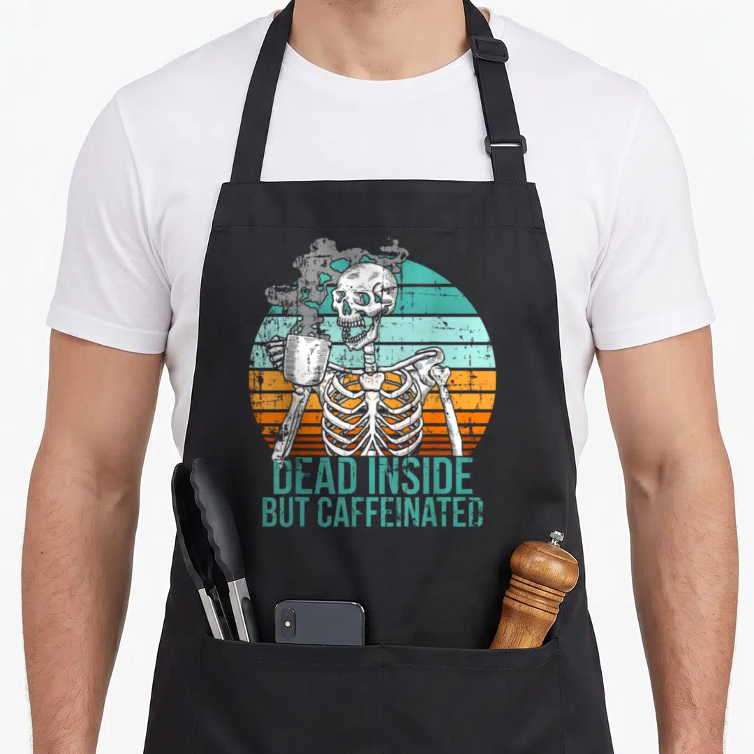 Dead coffee aprons - Skeleton holding mug in front of striped sun with teal text reading DEAD INSIDE BUT CAFFEINATED — shown on a black cotton canvas apron against a white background