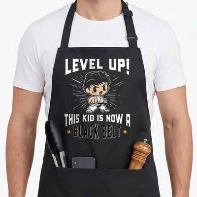 Karate uniform apron - Cartoon child in gi with LEVEL UP text and black belt award stars — shown on a black cotton canvas apron against a white background