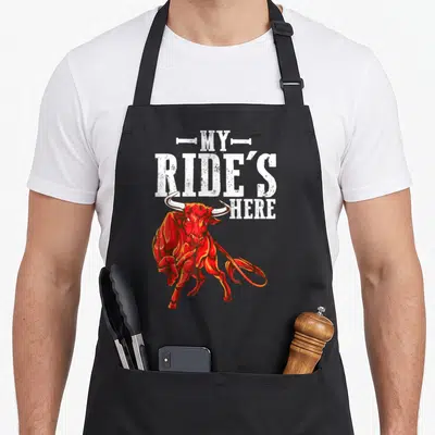Ride western aprons - Red cartoon bull below white stacked text reading MY RIDE'S HERE — shown on a black cotton canvas apron against a white background