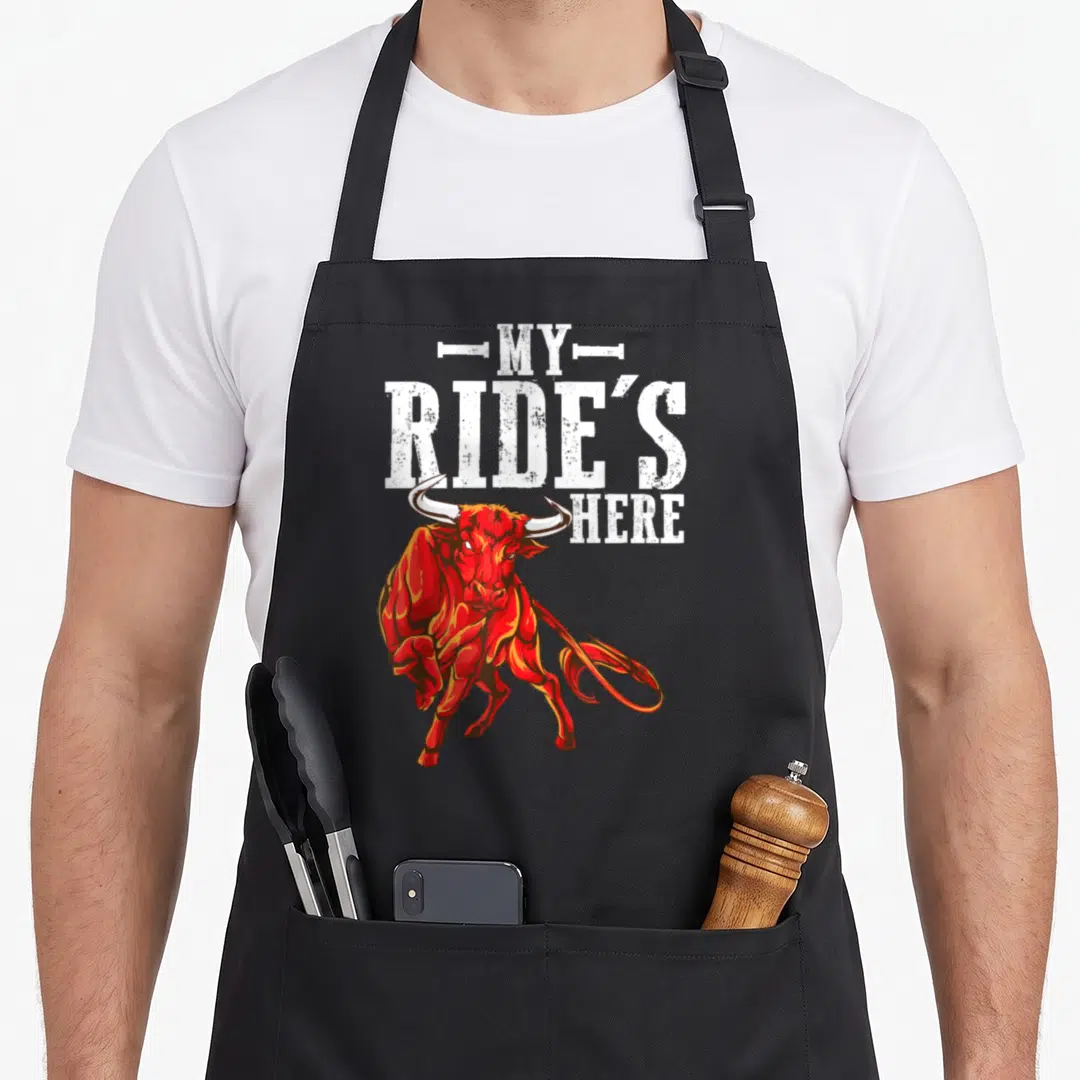 Ride western aprons - Red cartoon bull below white stacked text reading MY RIDE'S HERE — shown on a black cotton canvas apron against a white background