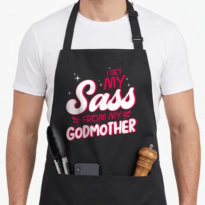 Godmother sass goddaughter apron - Stacked white and red text reading I get my sass from my godmother with sparkles — shown on a black cotton canvas apron against a white background
