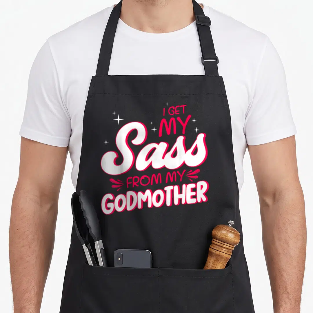 Godmother sass goddaughter apron - Stacked white and red text reading I get my sass from my godmother with sparkles — shown on a black cotton canvas apron against a white background