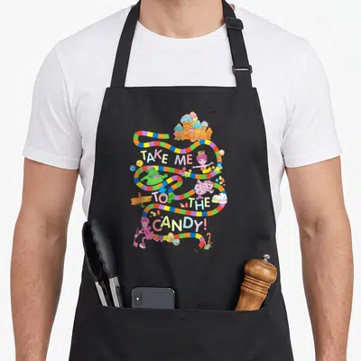 Funny land cotton candy apron - Multicolored winding path with TAKE ME TO THE CANDY text and cartoon sweets — shown on a black cotton canvas apron against a white background