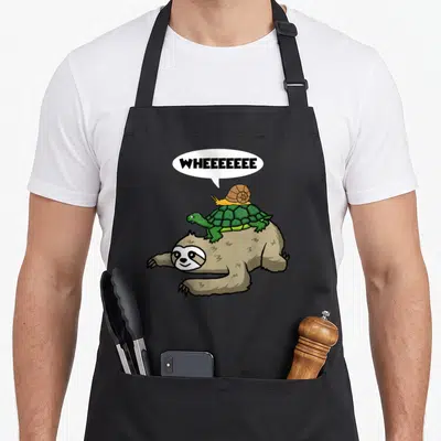 Snail sloth apron - Cartoon brown sloth carrying green turtle and yellow snail with WHEEEEEEEE text — shown on a black cotton canvas apron against a white background