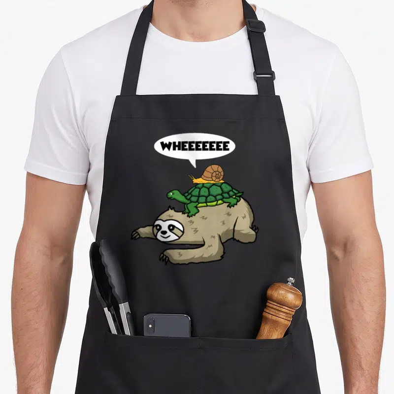 Snail sloth apron - Cartoon brown sloth carrying green turtle and yellow snail with WHEEEEEEEE text — shown on a black cotton canvas apron against a white background