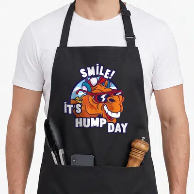 Smile camel apron - Cartoon orange camel with red sunglasses and SMILE HUMP DAY text in circular frame — shown on a black cotton canvas apron against a white background