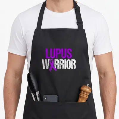 Lupus warrior ribbon apron - Purple and white distressed text LUPUS WARRIOR with ribbon replacing letter — shown on a black cotton canvas apron against a white background
