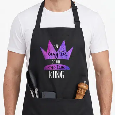 Daughter king apron - Purple blue watercolor crown with A daughter OF THE one true KING text — shown on a black cotton canvas apron against a white background