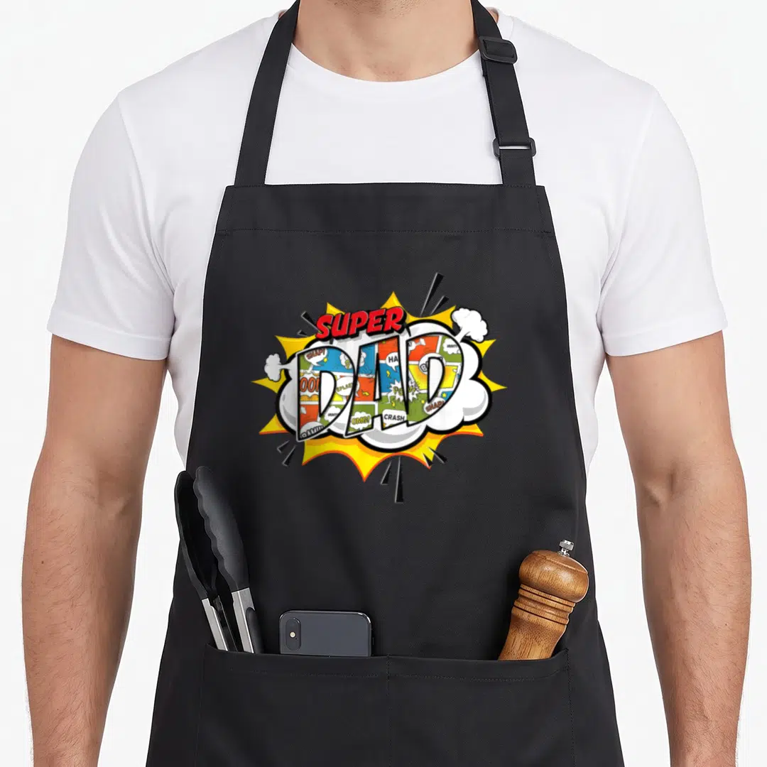 Comic robin dad apron - SUPER DAD text over yellow burst filled with comic panels and white smoke clouds — shown on a black cotton canvas apron against a white background