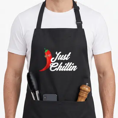 Chillin chili pepper apron - Red cartoon chili pepper wearing teal sunglasses next to white Just Chillin script text — shown on a black cotton canvas apron against a white background