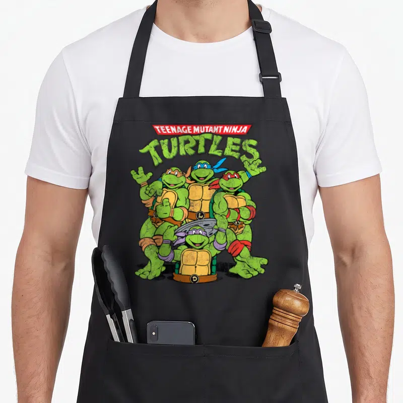 Funny teenage cooking aprons - Stacked TMNT logo with four turtles in group pose wearing colored masks — shown on a black cotton canvas apron against a white background