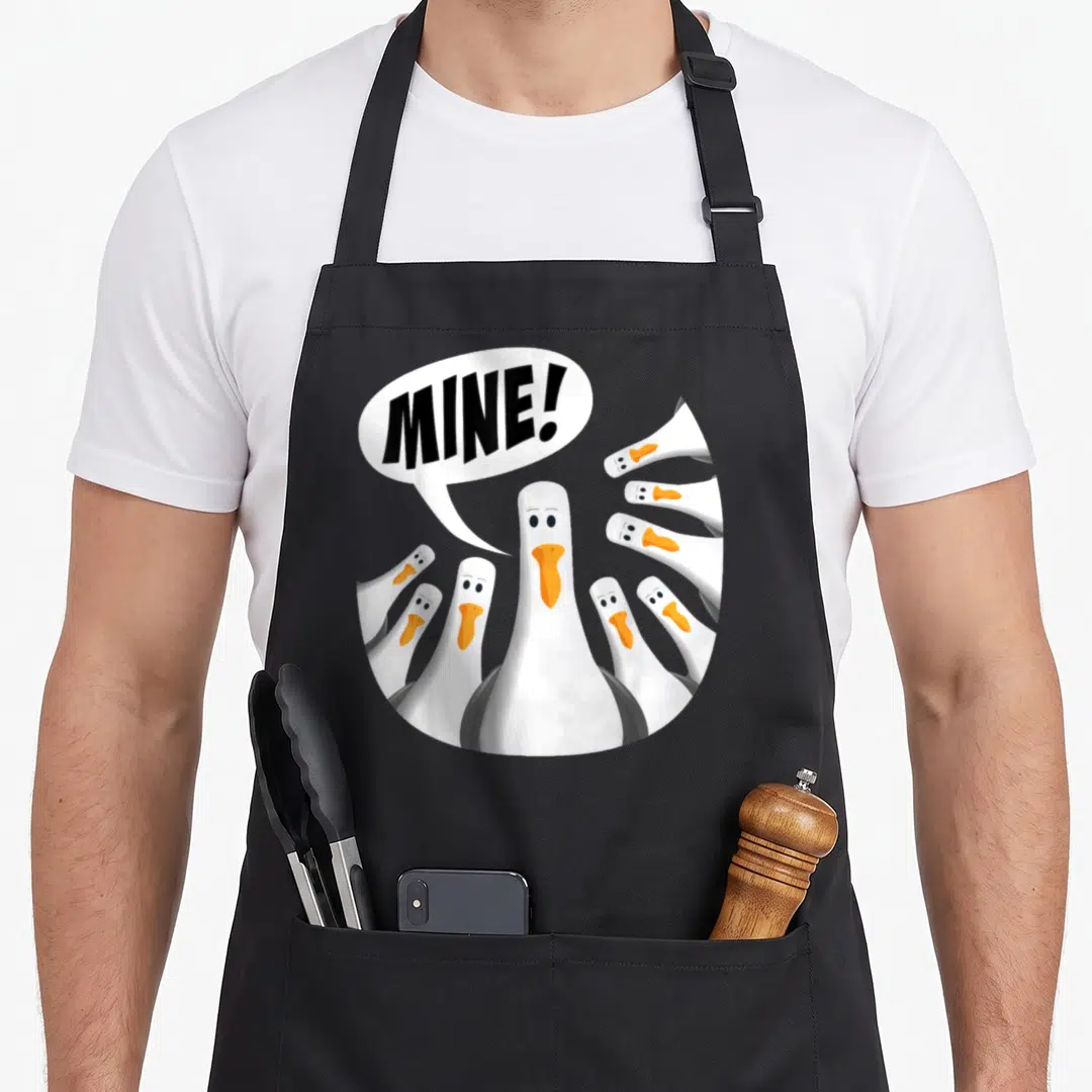 Mine seagull apron - Cartoon white goose with orange beak shouting MINE! surrounded by other geese heads — shown on a black cotton canvas apron against a white background