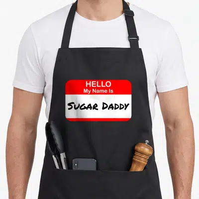 Hello daddy apron - Red and white name tag graphic with black handwritten text reading Hello My Name Is Sugar Daddy — shown on a black cotton canvas apron against a white background