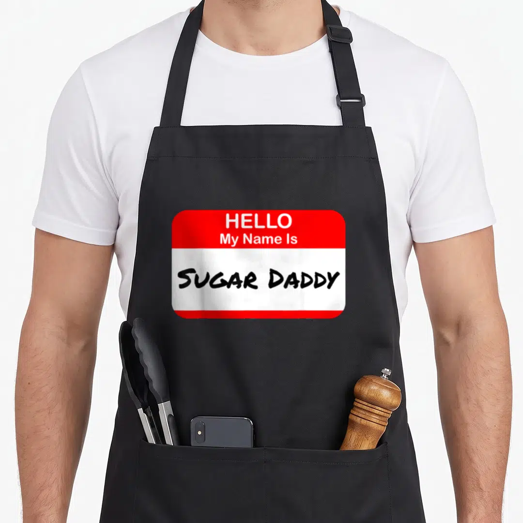 Hello daddy apron - Red and white name tag graphic with black handwritten text reading Hello My Name Is Sugar Daddy — shown on a black cotton canvas apron against a white background