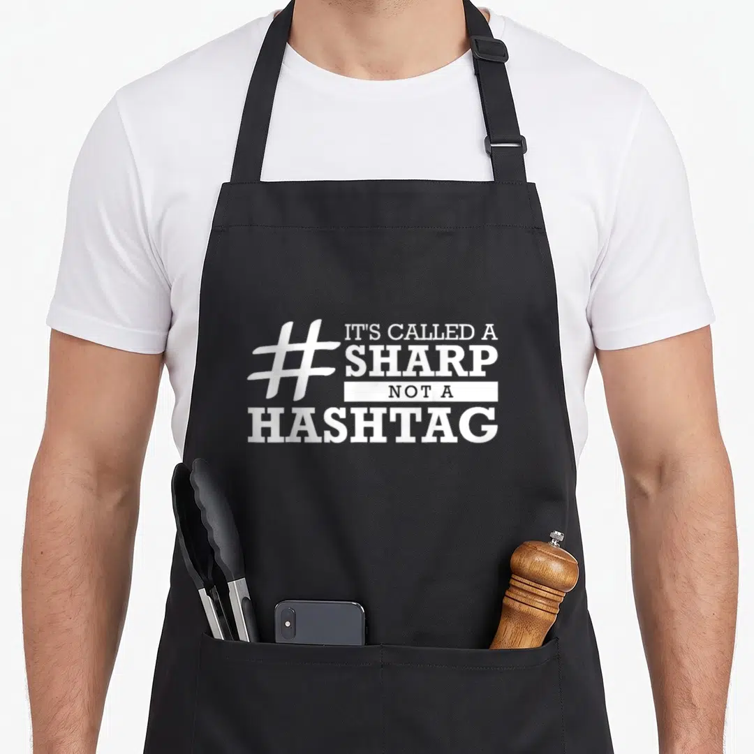 Hashtag band member apron - White sharp symbol and stacked text IT'S CALLED A SHARP NOT A HASHTAG in black and white — shown on a black cotton canvas apron against a white background