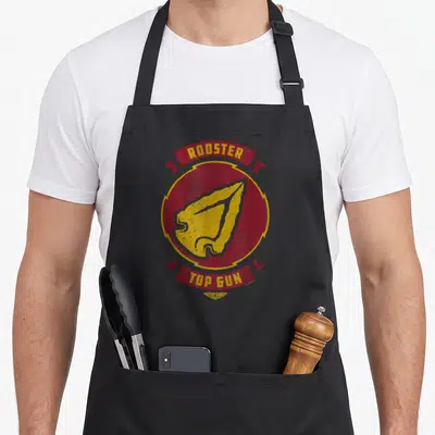 Gun rooster apron - Retro red circle with yellow rooster head and banner text reading ROOSTER and TOP GUN — shown on a black cotton canvas apron against a white background