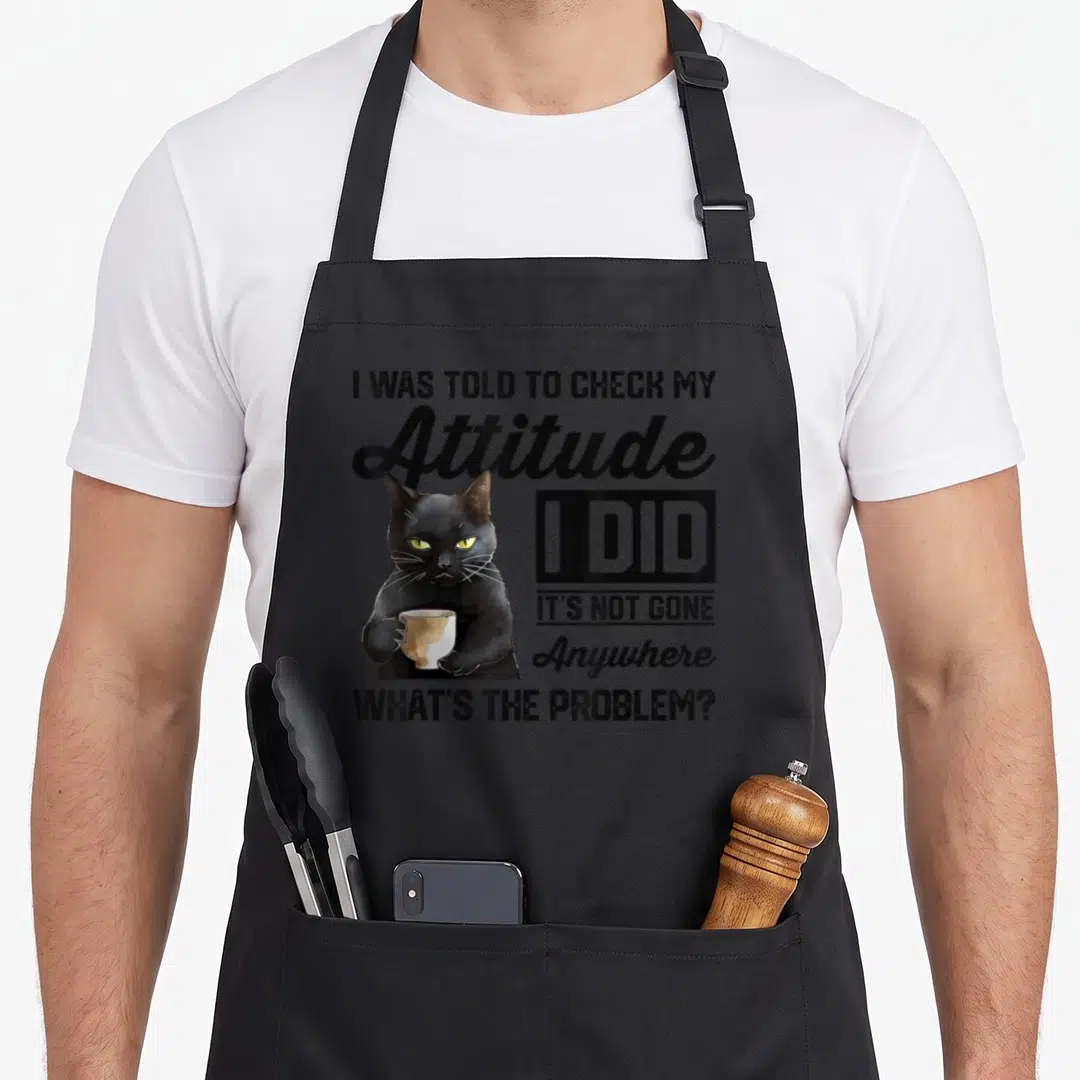 Check cat apron - Black cat holding stained cup with sarcastic text about attitude — shown on a black cotton canvas apron against a white background