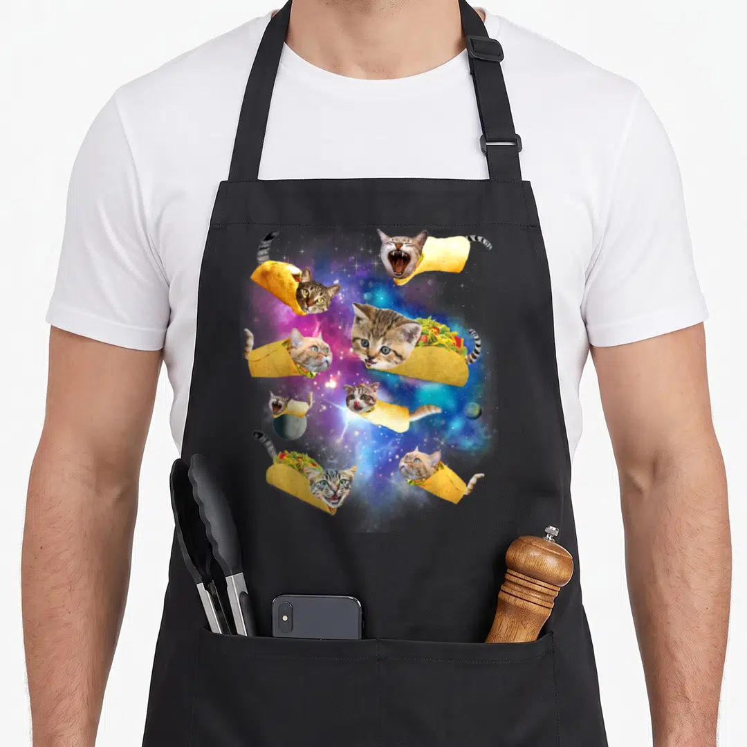 Burrito pizza cat apron - Six cats in taco shells floating among stars and two planets in a colorful nebula — shown on a black cotton canvas apron against a white background