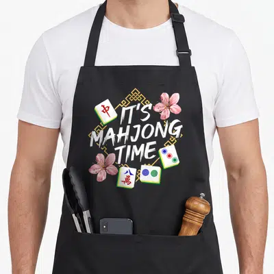 Chinese player mahjong apron - IT'S MAHJONG TIME text surrounded by four tiles and two pink flowers on a yellow — shown on a black cotton canvas apron against a white background