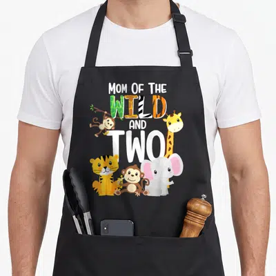 Theme safari birthday apron - MOM OF THE WILD AND TWO text with cartoon monkey, giraffe, tiger, and elephant in white — shown on a black cotton canvas apron against a white background