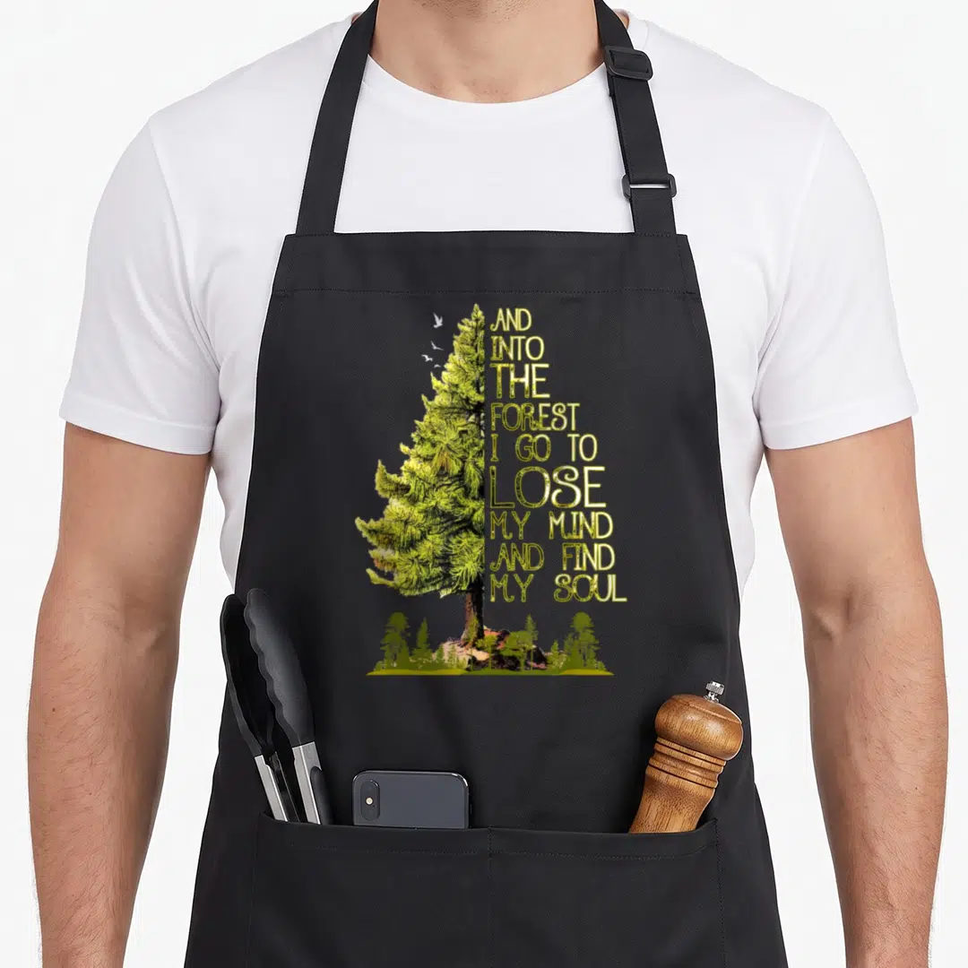 Forest find aprons - Realistic pine tree with stacked gold text reading And Into The Forest I Go To Lose My Mind — shown on a black cotton canvas apron against a white background
